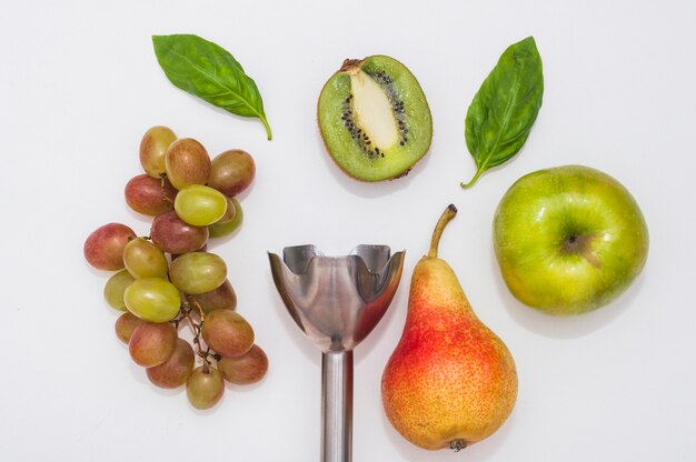 Uvas; albahaca; kiwi; Manzana y pera con batidora de mano eléctrica sobre fondo blanco