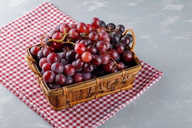 Uvas en una canasta sobre tela de picnic y yeso, vista de ángulo alto.