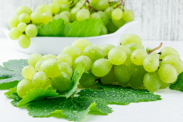 Uvas frescas con hojas en un plato sobre superficie blanca