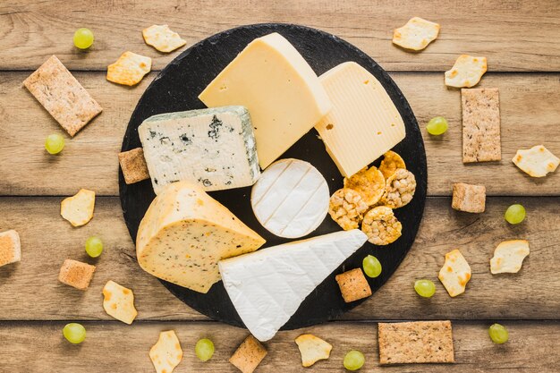 Uvas, galletas, pan crujiente y bloques de queso en pizarra redonda sobre el escritorio de madera