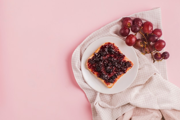 Uvas y rebanada de pan rojas con el atasco en la placa blanca sobre el mantel contra el contexto rosado