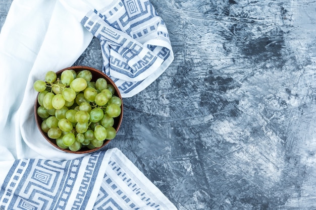 Uvas verdes en un cuenco de barro sobre yeso sucio y fondo de toalla de cocina. endecha plana.