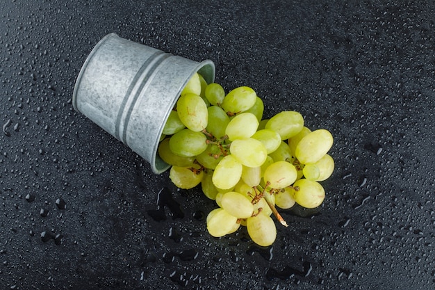 Uvas verdes esparcidas de un mini balde sobre un gris oscuro.