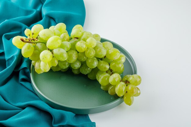 Uvas verdes en un plato sobre blanco y textil, vista de ángulo alto.