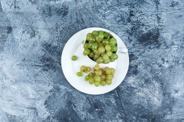 Uvas verdes en taza blanca y vista superior de la placa sobre un fondo de yeso sucio