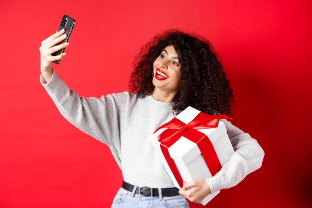 Vacaciones y concepto de tecnología. Mujer feliz tomando selfie con su regalo, sosteniendo presente y smartphone, de pie sobre fondo rojo.