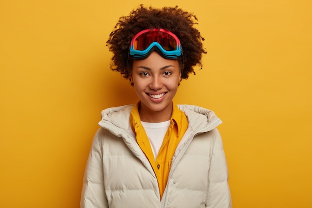 Vacaciones deportivas, estilo de vida de viaje y concepto de aventura de invierno. Alegre mujer africana con sonrisa dentuda, snowboard en las montañas, usa gafas de esquí y abrigo acolchado de plumas blancas