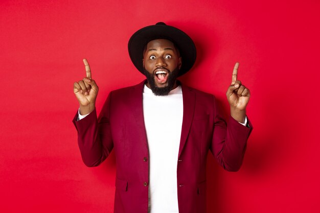 Vacaciones de invierno y concepto de compras. Alegre hombre afroamericano en traje de fiesta apuntando con el dedo hacia arriba, mostrando el logo y sonriendo feliz, fondo rojo.