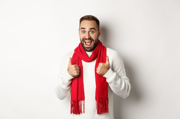 Vacaciones de invierno y concepto de compras. Hombre barbudo alegre como todo, mostrando los pulgares hacia arriba en señal de aprobación, de pie asombrado contra el fondo blanco.