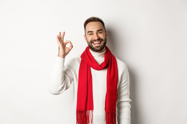 Vacaciones de invierno y concepto de compras. Hombre barbudo satisfecho riendo y mostrando signo bien, aprobar y como producto, de pie en bufanda y suéter de Navidad, fondo blanco.