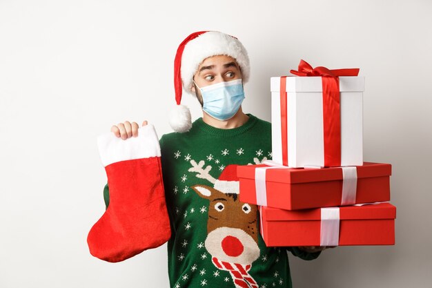 Vacaciones de invierno y concepto covid-19. Hombre feliz en mascarilla y gorro de Papá Noel trayendo regalos, sosteniendo un calcetín de Navidad y cajas de regalo, de pie sobre fondo blanco.