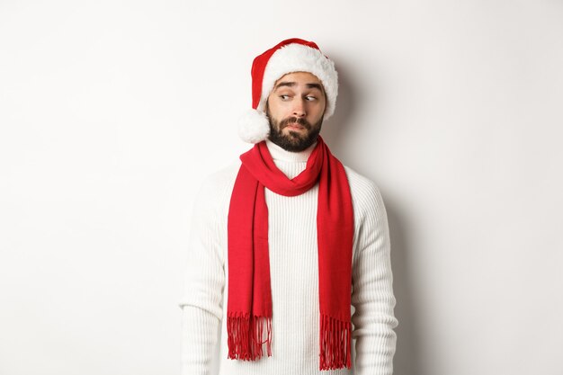 Vacaciones de invierno y concepto de fiesta de año nuevo. Chico curioso que mira a escondidas en la pancarta del logotipo de la izquierda, de pie con gorro de Papá Noel, celebrando la Navidad, de pie sobre fondo blanco