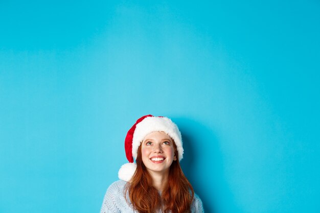 Vacaciones de invierno y concepto de Nochebuena. La cabeza de la linda chica pelirroja con gorro de Papá Noel, aparece desde la parte inferior y mira hacia arriba en el espacio de la copia, mirando el logotipo, de pie sobre un fondo azul.