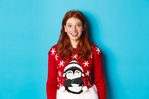 Vacaciones de invierno y concepto de Nochebuena. Chica pelirroja emocionada en suéter de Navidad mirando sorprendido a la cámara, de pie contra el fondo azul.