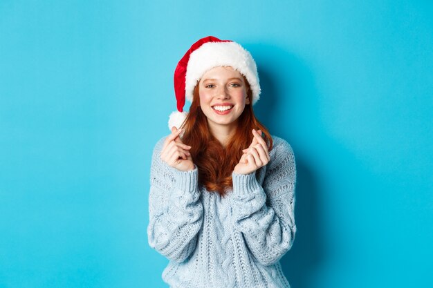 Vacaciones de invierno y concepto de Nochebuena. Chica pelirroja esperanzada con gorro de Papá Noel, pidiendo deseos en Navidad con los dedos cruzados, vistiendo gorro de Papá Noel, de pie sobre fondo azul.