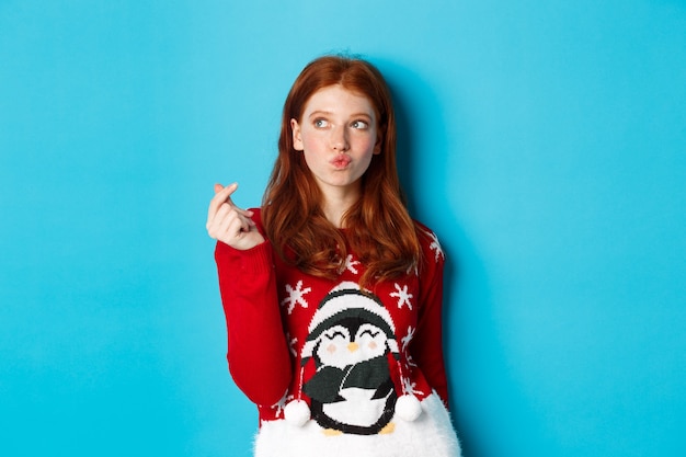 Vacaciones de invierno y concepto de Nochebuena. Encantadora mujer pelirroja en suéter de Navidad, mostrando el signo del corazón y pensando, mirando la esquina superior izquierda en el logo, fondo azul.