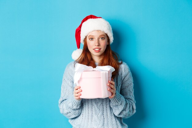 Vacaciones de invierno y concepto de Nochebuena. Linda chica pelirroja con gorro de Papá Noel, sosteniendo un regalo de año nuevo y mirando a cámara, de pie contra el fondo azul.