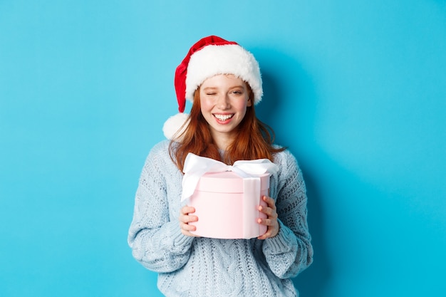 Vacaciones de invierno y concepto de Nochebuena. Linda chica pelirroja en suéter y gorro de Papá Noel, sosteniendo el regalo de año nuevo y mirando a la cámara, de pie contra el fondo azul