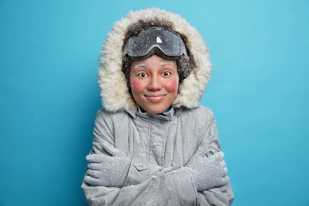 Vacaciones de invierno y concepto de recreación. Alegre mujer congelada siente frío después de ir a hacer snowboard en las montañas, tiembla y se abraza a sí misma para abrigarse, usa una chaqueta gris con guantes con capucha, se siente satisfecha