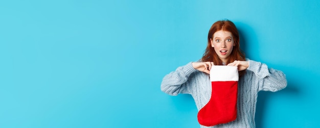 Vacaciones de invierno y concepto de regalos chica pelirroja divertida que parece sorprendida calcetín de navidad abierto y s