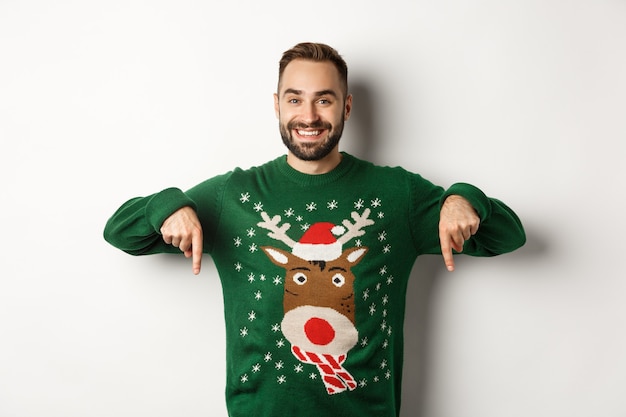 Vacaciones de invierno y navidad. Apuesto hombre sonriente con barba, apuntando con el dedo hacia abajo y mostrando una oferta promocional, vistiendo un suéter verde, de pie sobre fondo blanco.