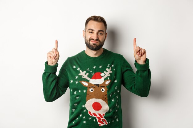 Vacaciones de invierno y navidad. Chico barbudo sin gracia en suéter divertido apuntando con el dedo hacia arriba, mostrando algo desagradable, fondo blanco.