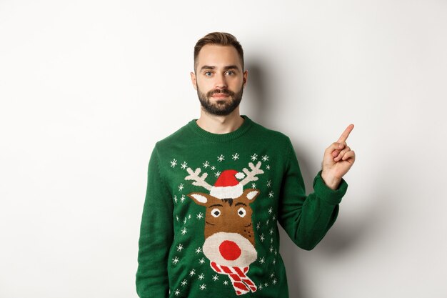 Vacaciones de invierno y navidad. Hombre guapo con barba en suéter verde apuntando con el dedo hacia la derecha, mostrando promo, de pie sobre fondo blanco.