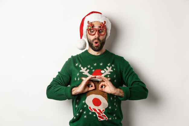 Vacaciones de Navidad, concepto de celebración. Chico feliz con gorro de Papá Noel y gafas de fiesta burlándose de suéter divertido, de pie sobre fondo blanco.