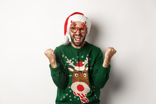 Vacaciones de Navidad, concepto de celebración. Hombre feliz con gorro de Papá Noel triunfando, vistiendo divertidas gafas de fiesta y regocijándose, de pie sobre fondo blanco.