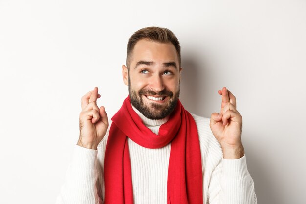 Vacaciones navideñas. Hombre atractivo feliz pidiendo deseos en las vacaciones de invierno, cruzar los dedos para la buena suerte, mirando hacia arriba, de pie sobre fondo blanco.