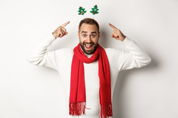 Vacaciones navideñas. Hombre emocionado celebrando las vacaciones de invierno, vistiendo accesorios de fiesta de año nuevo y pañuelo rojo, de pie contra el fondo blanco.