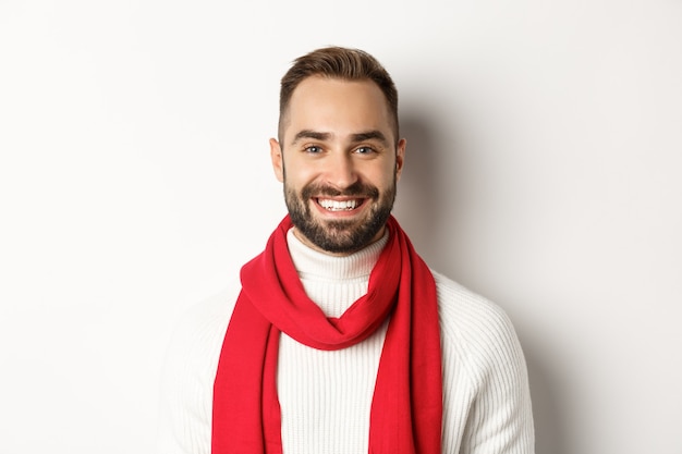 Vacaciones navideñas. Hombre guapo con barba en bufanda roja y suéter sonriendo, fondo blanco.