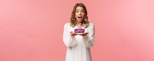 Vacaciones primavera y concepto de fiesta Feliz alegre guapa mujer rubia celebrando cumpleaños sosteniendo un pedazo de pastel con una vela encendida haciendo que el deseo se vea divertido de pie fondo rosa