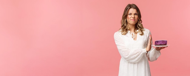 Vacaciones de primavera y concepto de fiesta Retrato de una chica rubia insatisfecha y disgustada con vestido blanco que rechaza rechazar comer pastel insípido haciendo muecas y muestra la señal de stop en el fondo rosa del postre