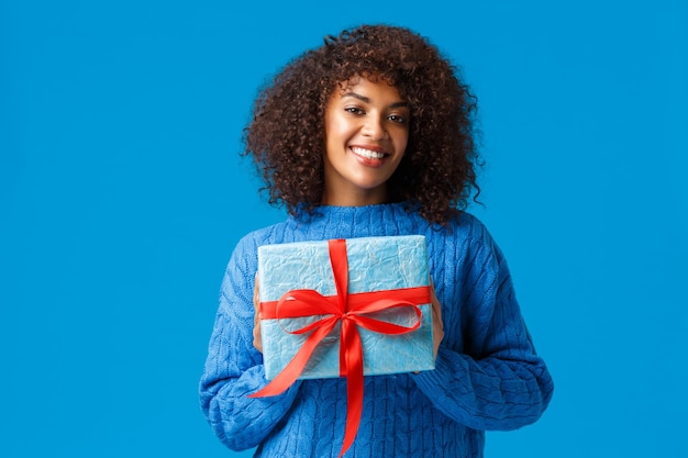 Vacaciones, temporada de invierno y concepto de Navidad. Encantadora linda mujer afroamericana envolvió su regalo para darle a su novio el día de San Valentín, sonriendo alegre, diciendo feliz año nuevo.