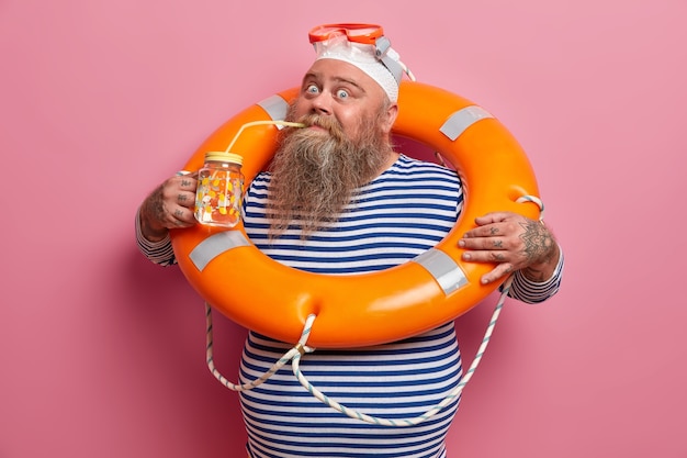 Vacaciones de verano y concepto de recreación. Un hombre adulto barbudo regordete bebe agua fresca durante un día caluroso, posa con una boya naranja de rescate, usa gorro de baño y gafas, aislado en la pared rosa.