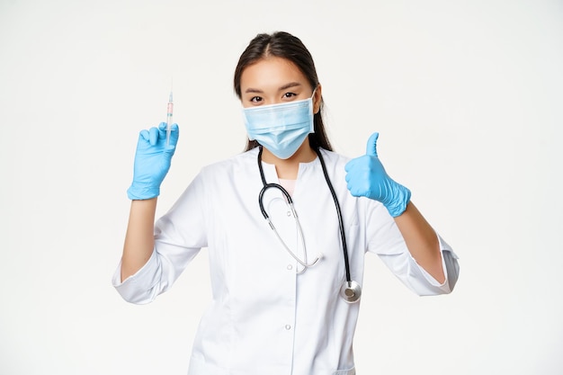 Vacunación y asistencia sanitaria Covid-19. Doctora asiática en mascarilla médica y guantes, sosteniendo la jeringa de la vacuna y mostrando el pulgar hacia arriba, fondo blanco.