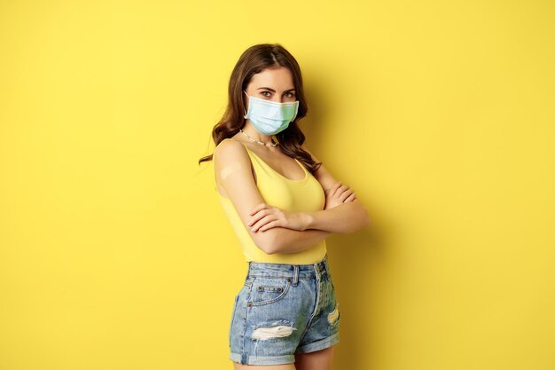 Vacunación covid-19. Hermosa mujer joven con máscara médica facial fue vacunada durante la pandemia, de pie sobre fondo amarillo.