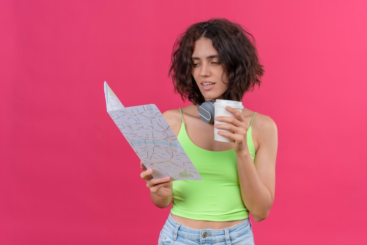 Foto gratuito una valiente mujer joven y bonita con el pelo corto en la parte superior de la cosecha verde en auriculares sosteniendo una taza de café de plástico mirando un mapa con confianza