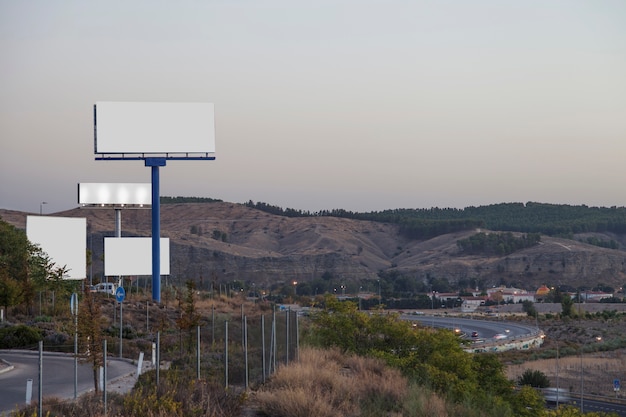 Vallas publicitarias blancas en carretera con montañas al fondo.