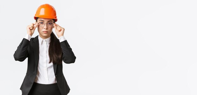 Vamos a trabajar Serio, seguro de sí mismo, el ingeniero jefe asiático femenino inspecciona la empresa con casco de seguridad y traje puesto en gafas de fondo blanco listo para trabajar