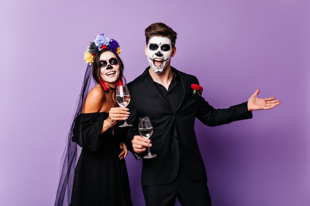 Vampiros felices bebiendo vino sobre fondo morado. Foto de estudio de pareja en atuendos tradicionales de zombies mexicanos.