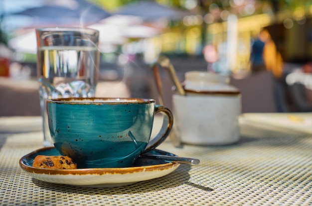 El vapor se eleva sobre una taza de café expreso, un vaso de agua y galletas en la mesa, la luz del sol y el enfoque selectivo, el espacio vacío, la publicidad o el desayuno del café.