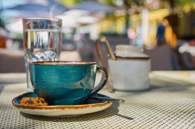 El vapor se eleva sobre una taza de café expreso, un vaso de agua y galletas en la mesa, la luz del sol y el enfoque selectivo, el espacio vacío, la publicidad o el desayuno del café.