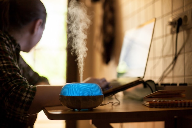 Foto gratuita vapor que sale del difusor de aceite esencial con led azul mientras la mujer trabaja en la computadora portátil.