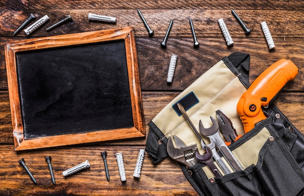 Varias herramientas en toolbag cerca de pizarra en blanco y pernos sobre fondo de madera