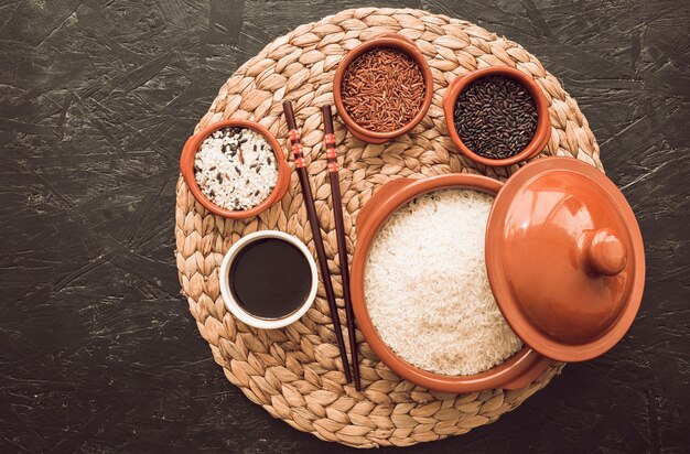 Variedad de granos de arroz sin cocinar diferentes en un tazón con salsa de soja sobre el mantel individual