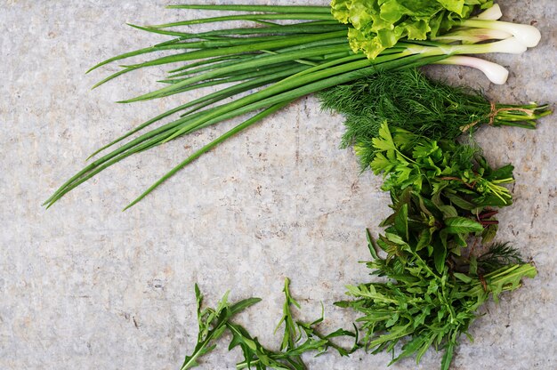 Variedad de hierbas orgánicas frescas (lechuga, rúcula, eneldo, menta, lechuga roja y cebolla) sobre fondo gris en estilo rústico. Vista superior