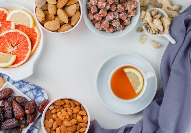 Variedad de nueces con una taza de té, dátiles, rodajas de cítricos y jengibre en platos blancos.