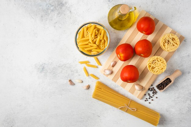 Variedad de pasta cruda, botella de aceite de oliva, granos de pimienta y tomates sobre fondo blanco.